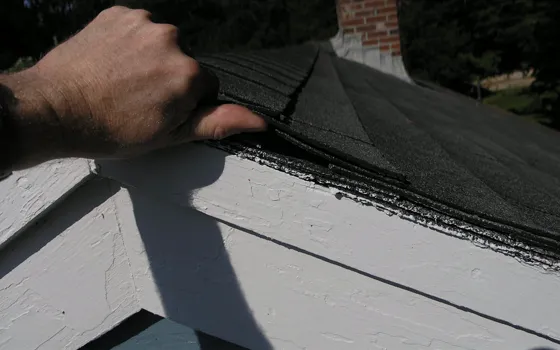 Example photo: A hand lifts the shingle edge at the fascia, showing stacked layers of roofing